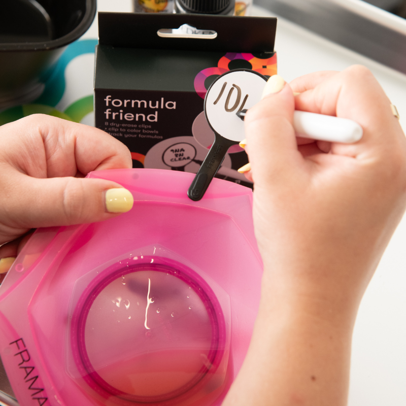 Stylist writing a colour formula onto a Framar Formula Friend clip attached to a pink mixing bowl using the dry erase marker